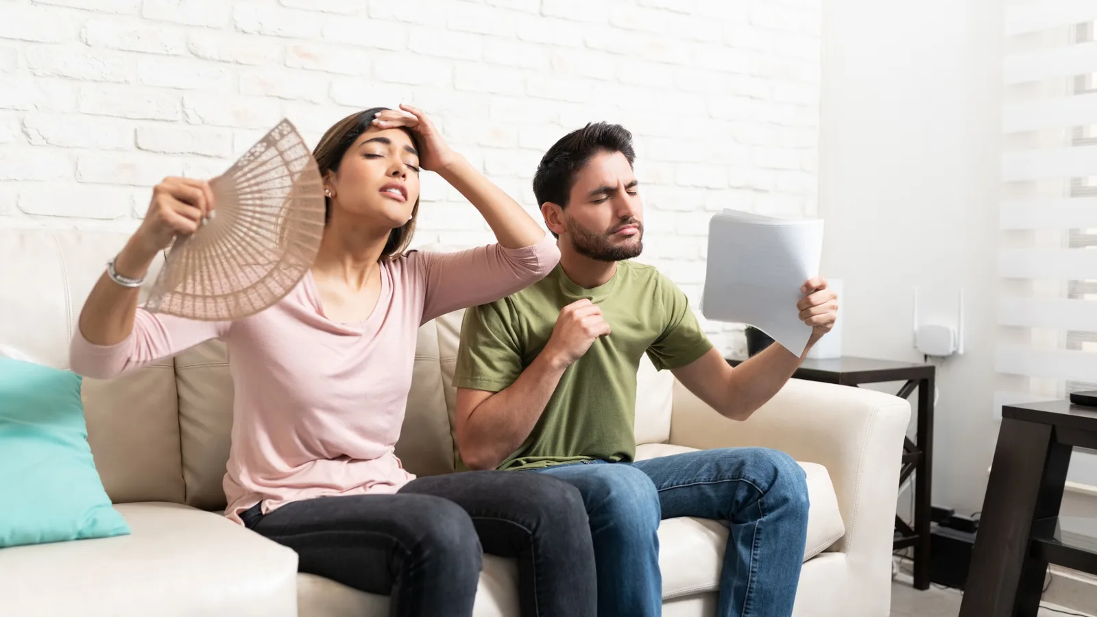 Couple feeling hot indoors, woman using a fan and man fanning himself with papers, emphasizing the need for effective AC maintenance during summer.