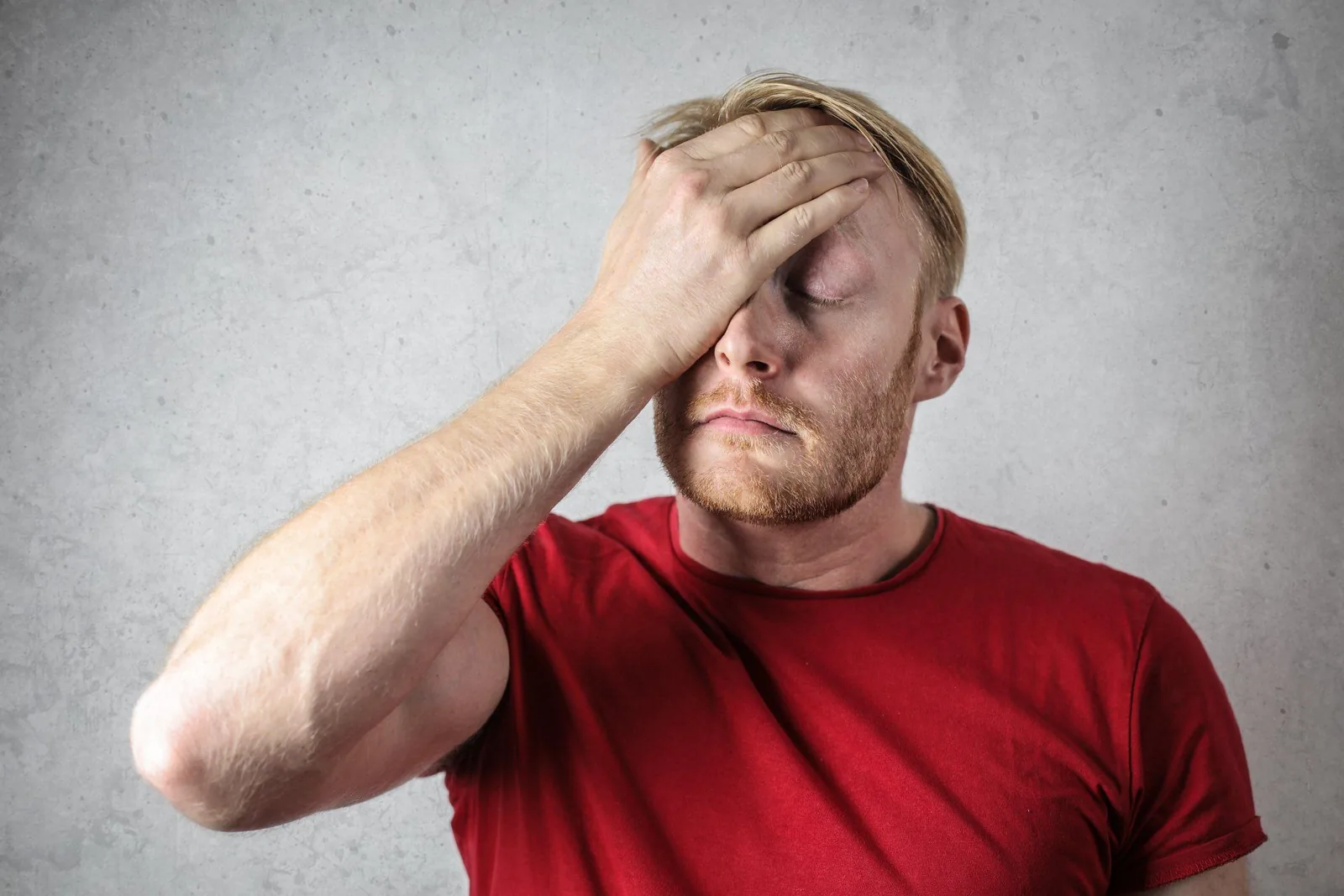 Man in a red shirt expressing frustration or concern, touching his forehead, against a textured gray background
