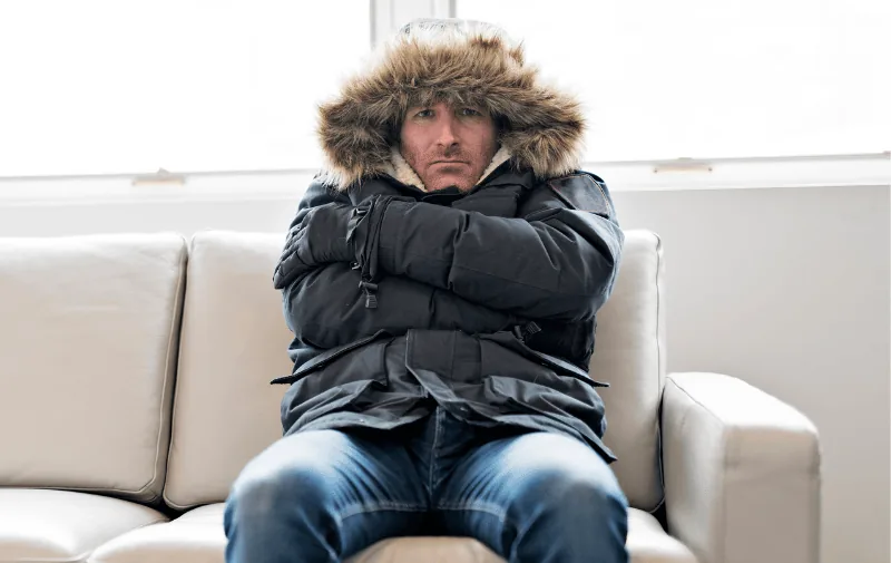 Man sitting on a couch, bundled in a fur-lined winter coat, looking cold and uncomfortable, illustrating heating issues in a home.