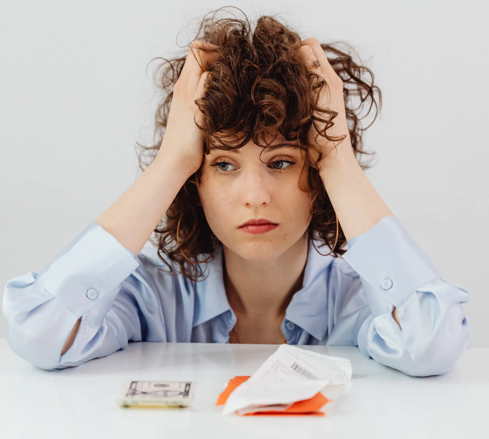 Woman with curly hair looking stressed, holding her head, with money and receipts on a table, reflecting concerns about