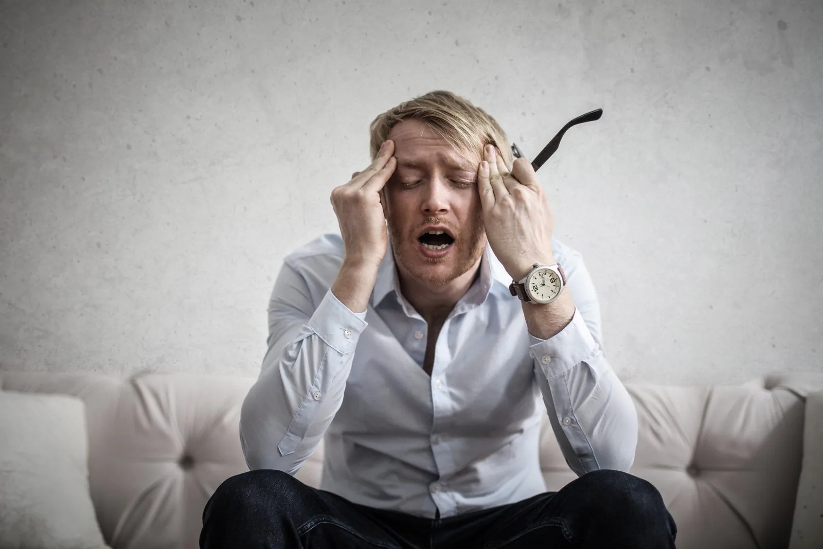 Frustrated man in a white shirt sitting on a couch, holding his head, conveying stress likely related to furnace issues or HVAC problems.