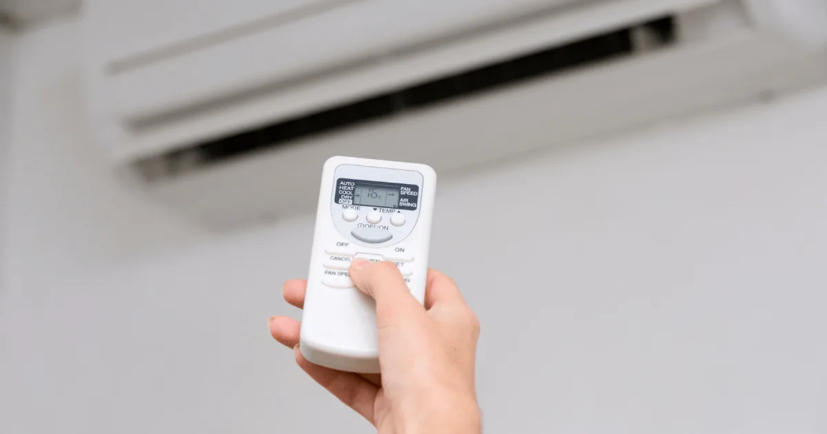 Hand holding air conditioner remote control, adjusting settings for optimal comfort and energy efficiency.