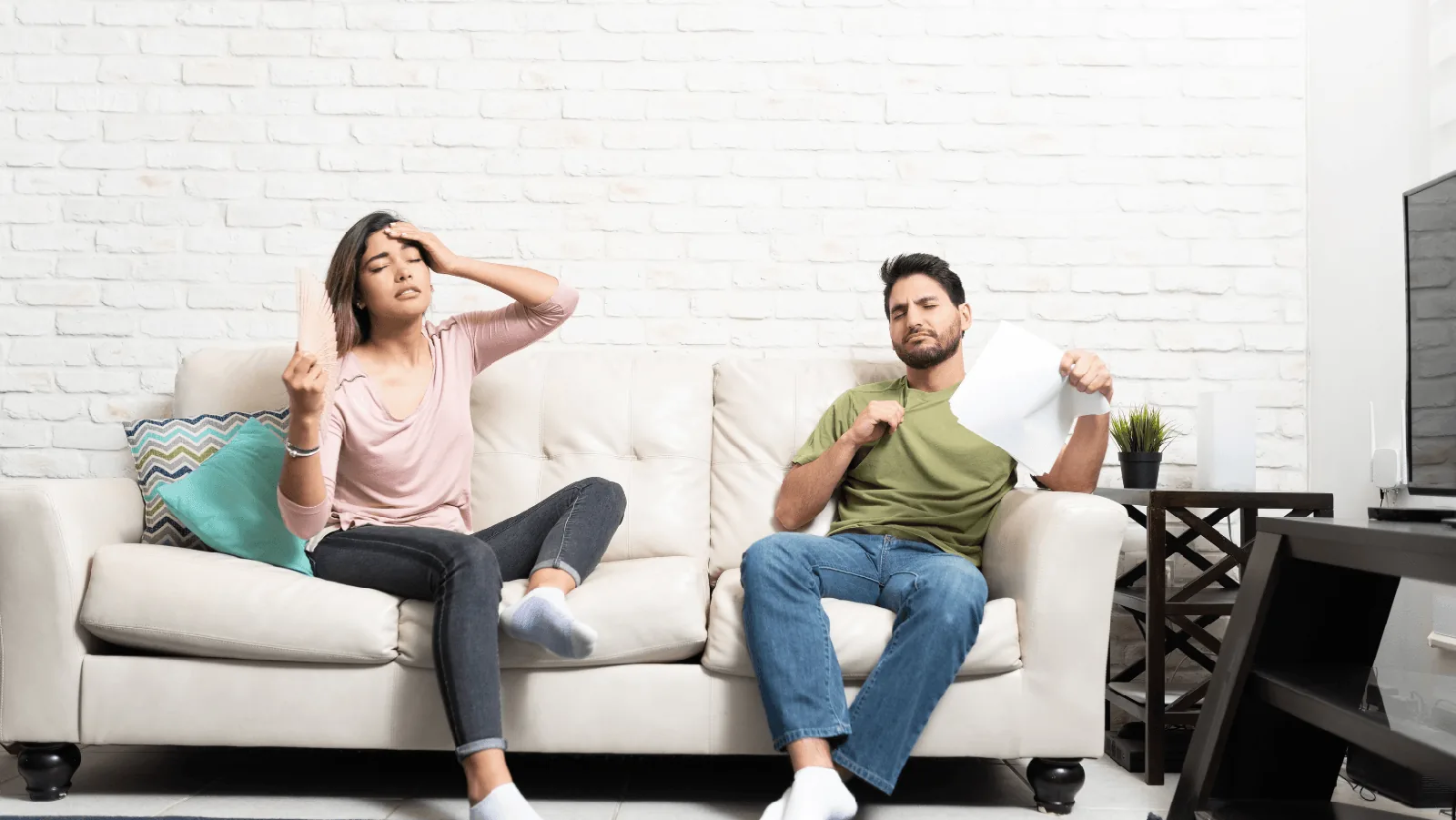 Couple sitting on a couch indoors, woman fanning herself looking uncomfortable and man holding a piece of paper