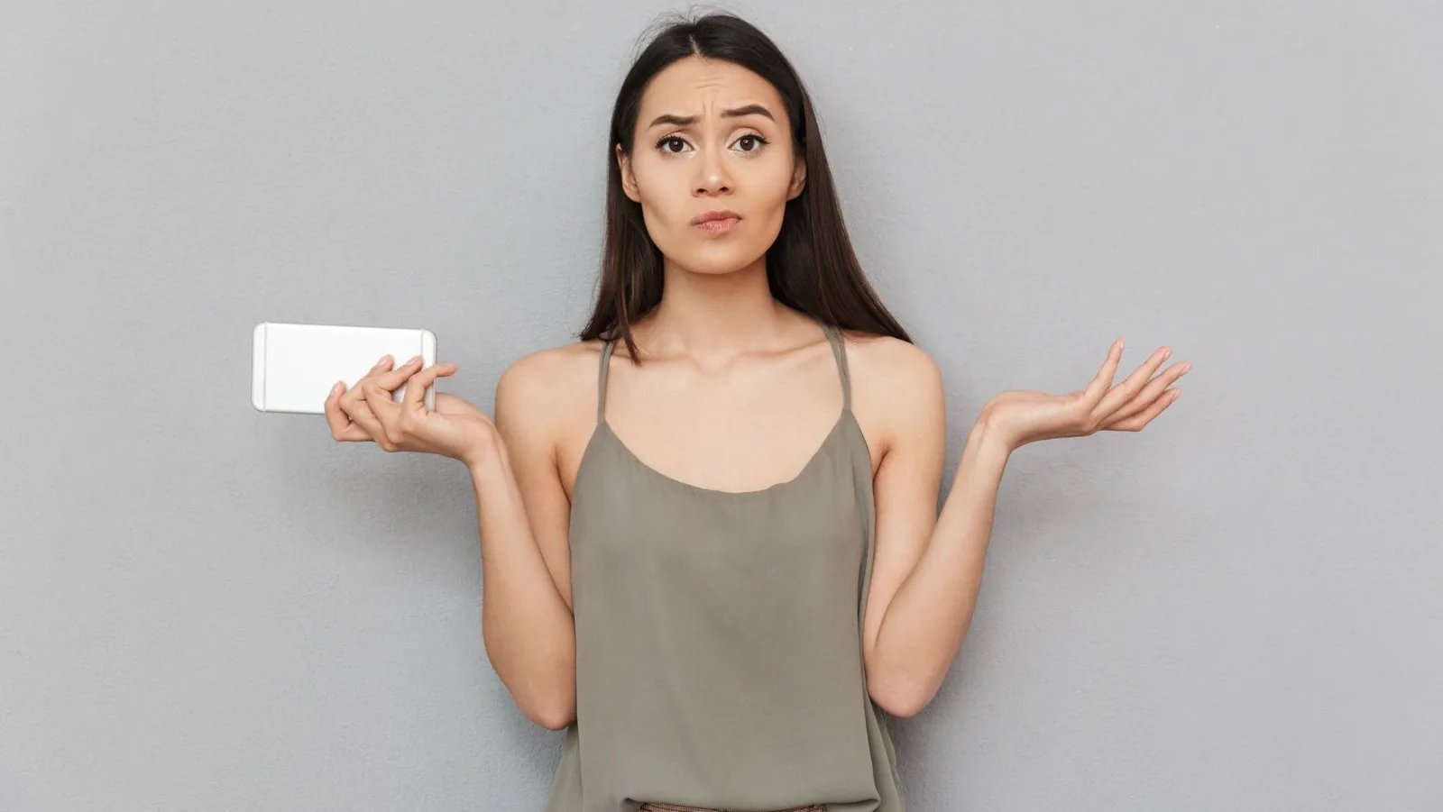 Confused woman holding a smartphone against a gray background, representing inquiries about HVAC maintenance and services.