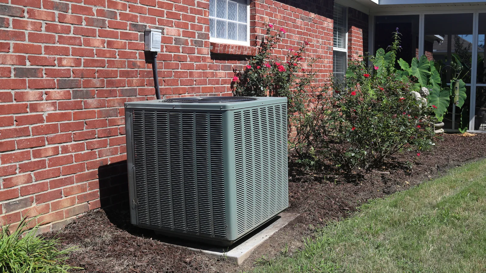 Outdoor mini split HVAC unit beside brick wall and flowering shrubs, illustrating residential heating and cooling solutions.