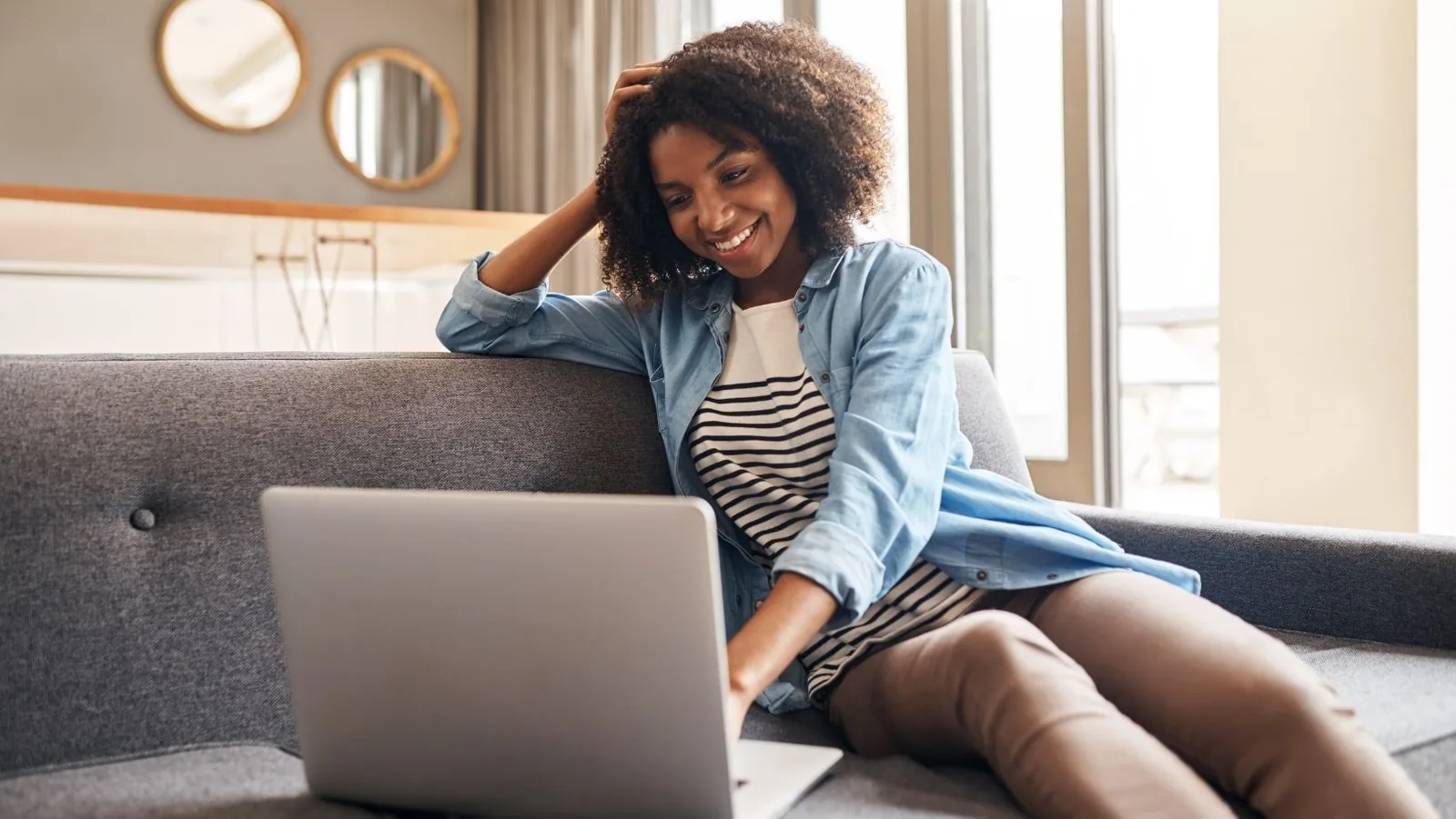 Person smiling while sitting on a couch, using a laptop, in a modern living room setting, reflecting comfort and engagement in home activities.