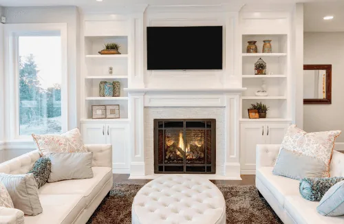 Cozy living room featuring white sofas, a circular tufted ottoman, a modern fireplace, and built-in shelves, emphasizing
