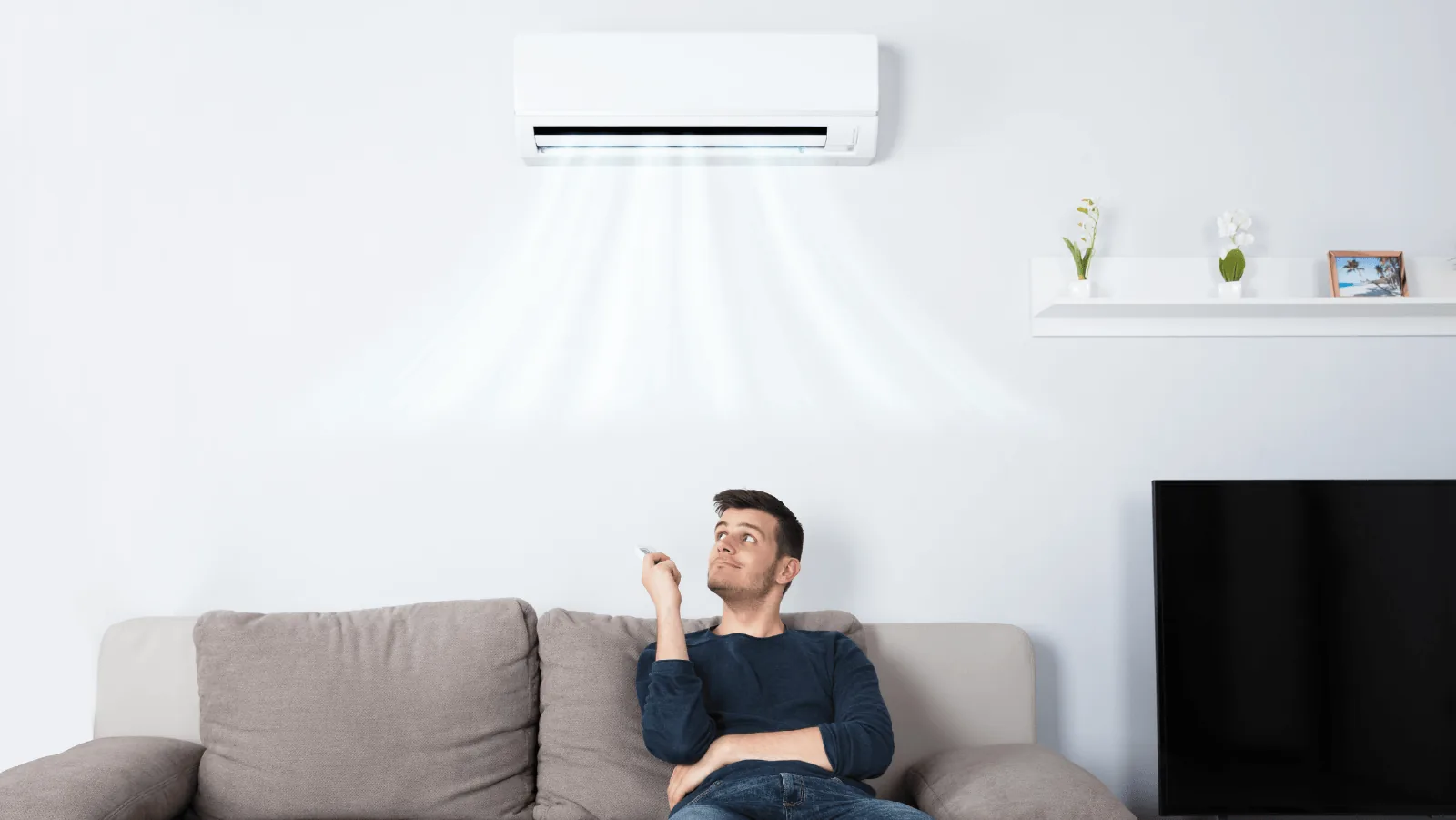 Man sitting on a couch using a remote control to adjust a wall-mounted ductless heat pump, emphasizing indoor air quality and HVAC services.