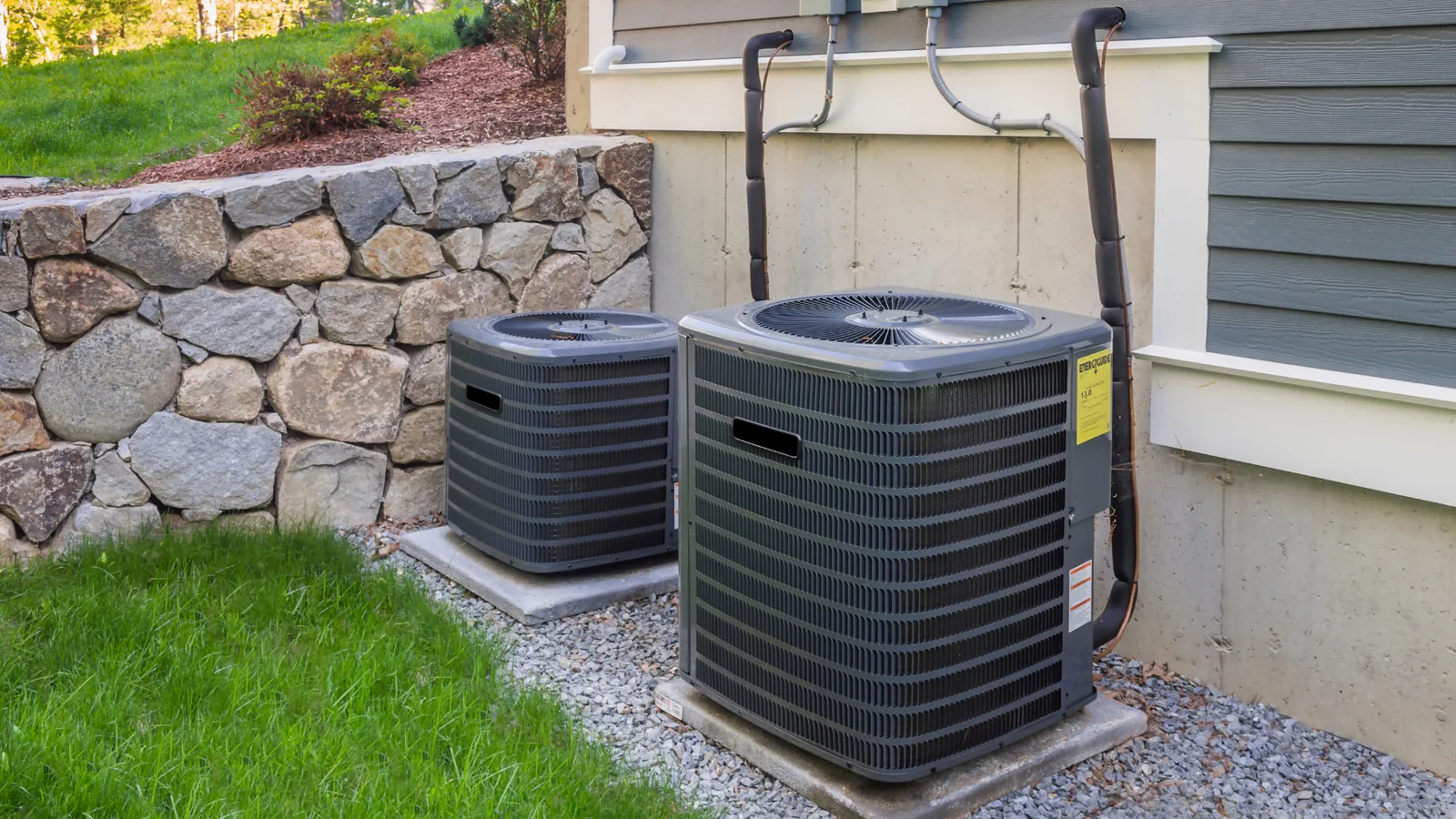 HVAC units installed outside a home, showcasing two air conditioning compressors on concrete pads, surrounded by a stone