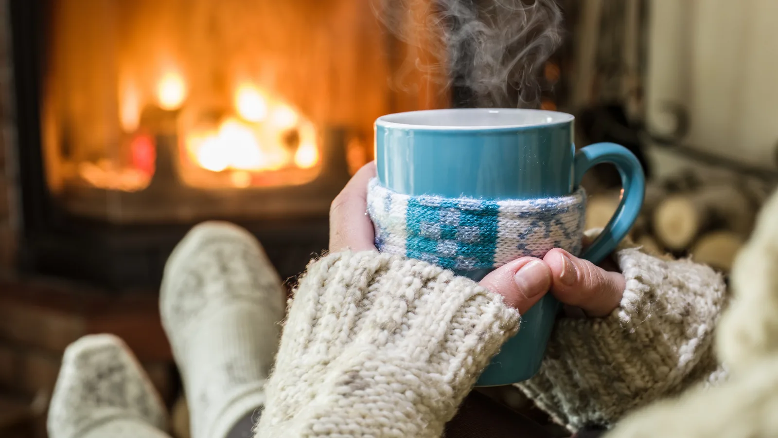 Cozy scene of a person holding a steaming mug in front of a fireplace, wearing a warm sweater, symbolizing comfort and warmth during winter months.