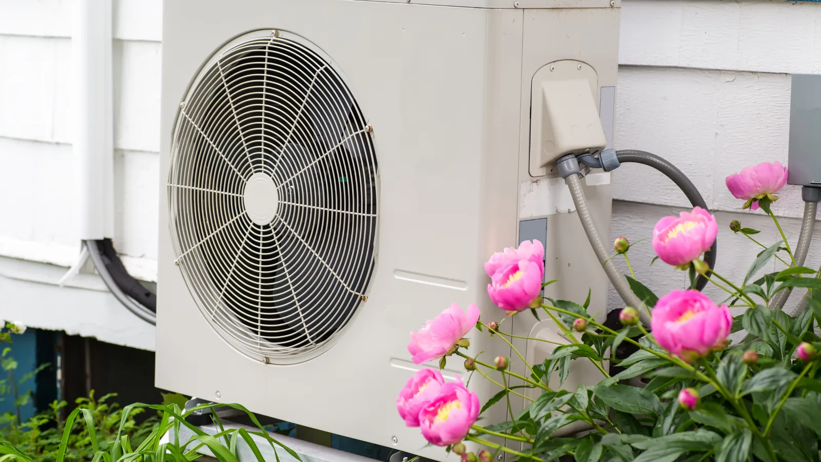 Ductless mini split air conditioning unit installed outside a home, surrounded by pink peony flowers, illustrating HVAC