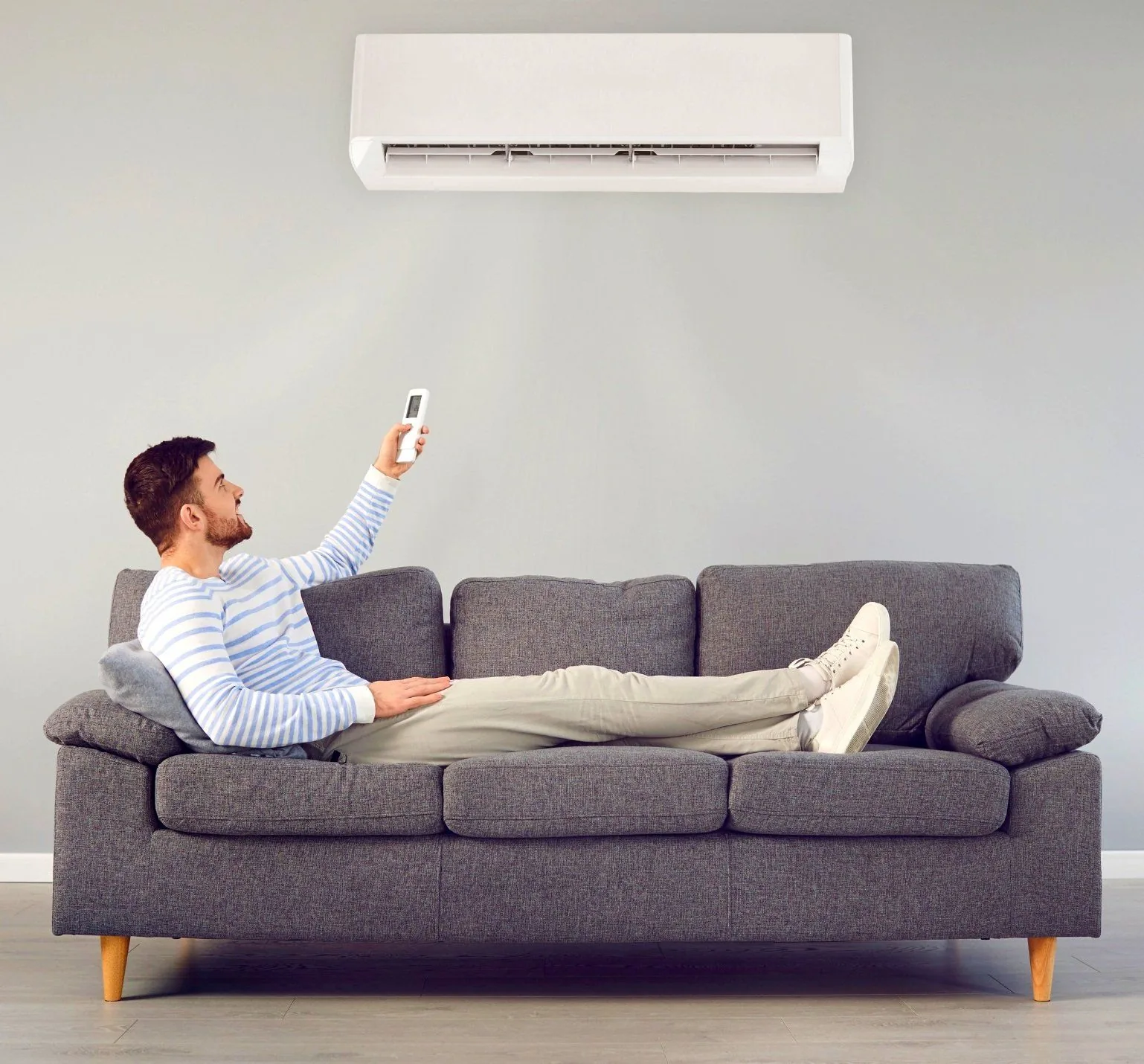 Man relaxing on a gray couch, using a remote control to adjust an air conditioning unit mounted on the wall