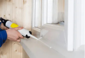 Person using a caulking gun to seal cracks around a window, emphasizing the importance of sealing air leakages for HVAC efficiency and indoor comfort.